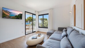 A modern living room with a large grey sofa, a white ottoman, and a flat-screen TV displaying a scenic landscape. - APARTAMENTO ORATGE 2 (Puerto Pollensa)