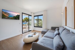 A modern living room with a large grey sofa, a white ottoman, and a flat-screen TV displaying a scenic landscape. - APARTAMENTO ORATGE 2 (Puerto Pollensa)
