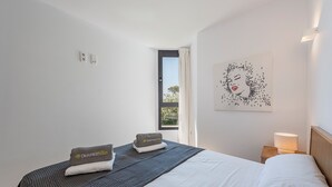 A hotel room with a bed, a nightstand, a lamp, a window with curtains, and a wall art piece. - APARTAMENTO ORATGE 2 (Puerto Pollensa)