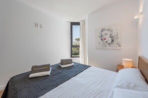 A hotel room with a bed, a nightstand, a lamp, a window with curtains, and a wall art piece. - APARTAMENTO ORATGE 2 (Puerto Pollensa)