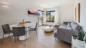 A modern living room with a dining area, a grey sofa, a round coffee table, and a large window with a view. - APARTAMENTO ORATGE 2 (Puerto Pollensa)