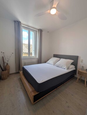 Ein Schlafzimmer mit Bett, Deckenventilator, Fenster mit Vorhängen und einem kleinen Nachttisch.