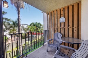 A balcony with a wooden door, a table, two chairs, and a view of a fenced yard with palm trees.