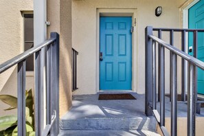 A blue door with a metal staircase leading up to it, a doormat, and a wall-mounted light fixture.