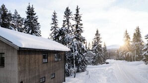 A wooden cabin with a snow-covered roof, surrounded by evergreen trees and a snow-covered driveway. - Gamla Wallesvägen 10 - Bruksvallarna (Bruksvallarna)