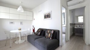 Una sala de estar moderna con un sofá gris, una mesa de comedor blanca con sillas, una lámpara colgante blanca y una cocina visible a través de un puerta abierta.
