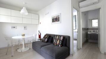 Ein modernes Wohnzimmer mit einem grauen Sofa, einem weißen Esstisch mit Stühlen, einer weißen Pendelleuchte und einem durch eine offene Tür einsehbaren Küchenbereich.