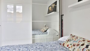 Un dormitorio con cama, armario, estante con un cuadro y ventana.
