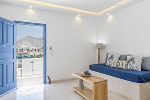 Une chambre moderne avec un lit, une petite table, une lampe et une vue sur des immeubles et une montagne, visible à travers une porte ouverte.