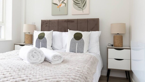 A neatly made bed with rolled towels, a brown headboard, two bedside tables with lamps, and two wall art pieces. - Sapphire Court Flat 69 2-Bedroom for family corporate or business travel (Birmingham)