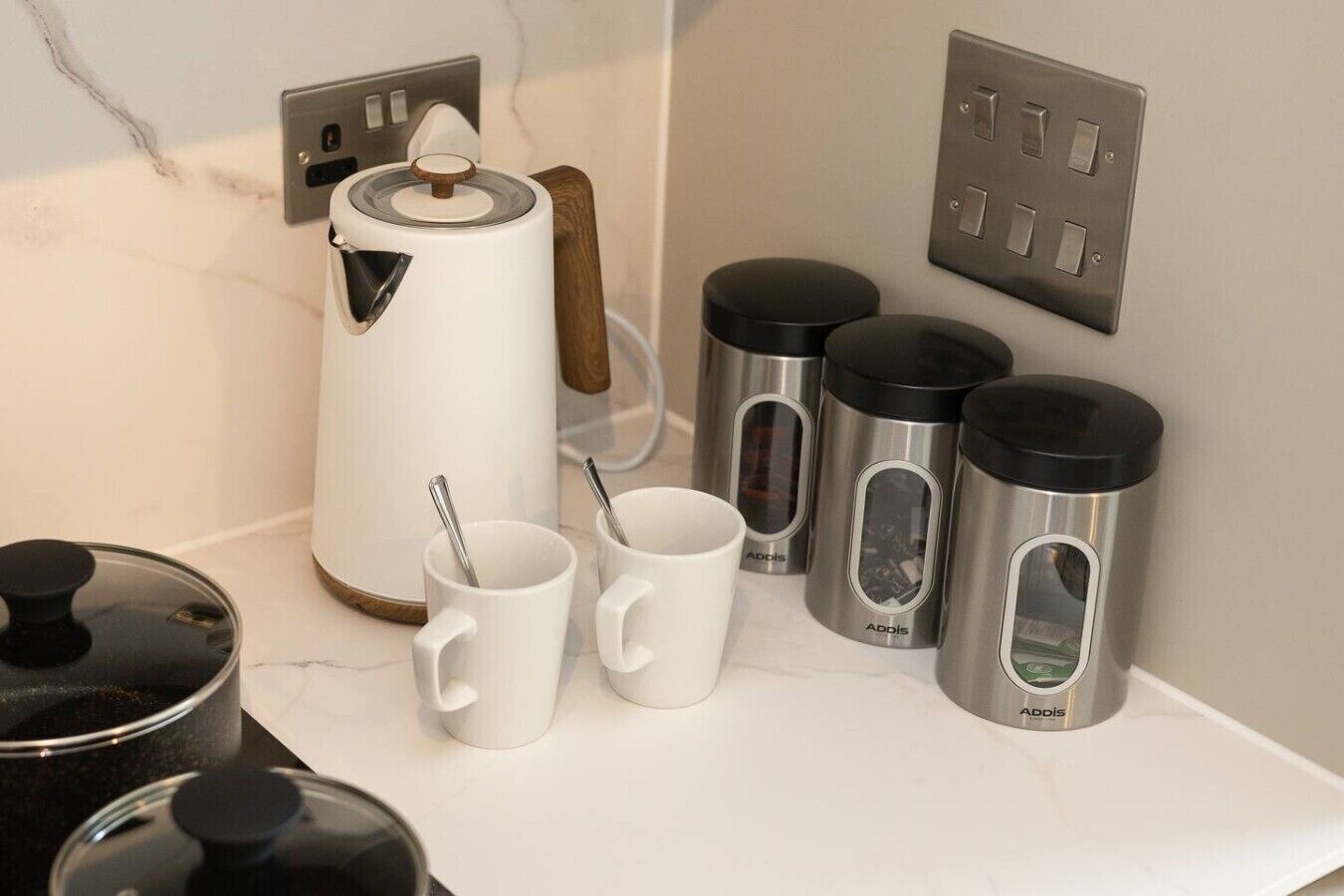 Una encimera de cocina con una cafetera, dos tazas blancas y cuatro termos de acero inoxidable.