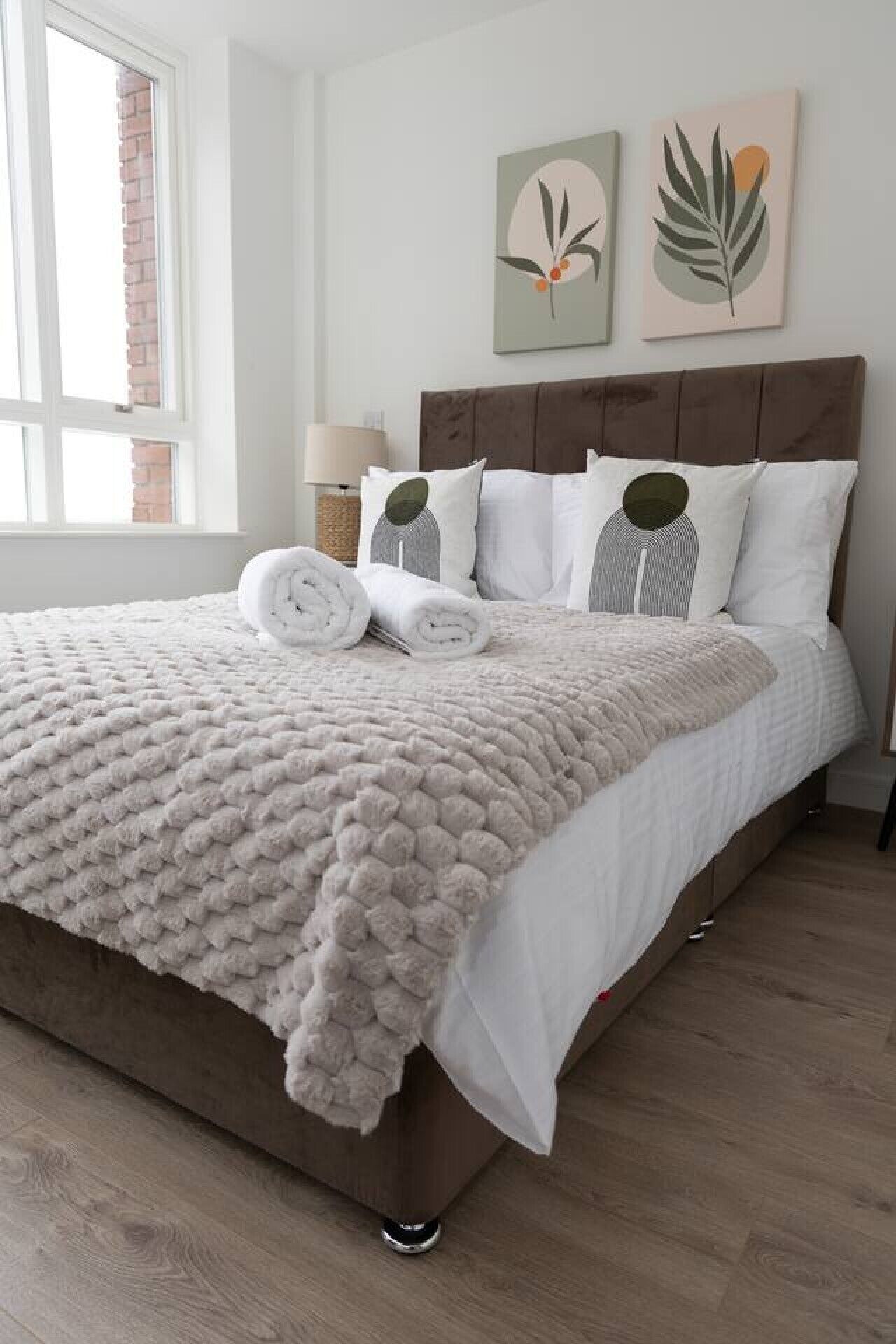 A neatly made bed with a fluffy white blanket and rolled towels, a brown headboard, and two wall-mounted artworks.