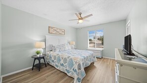 Ein Schlafzimmer mit einem Bett, einem Deckenventilator, einem Fenster mit Jalousien, einem Schreibtisch mit Computer und einer kleinen Pflanze.