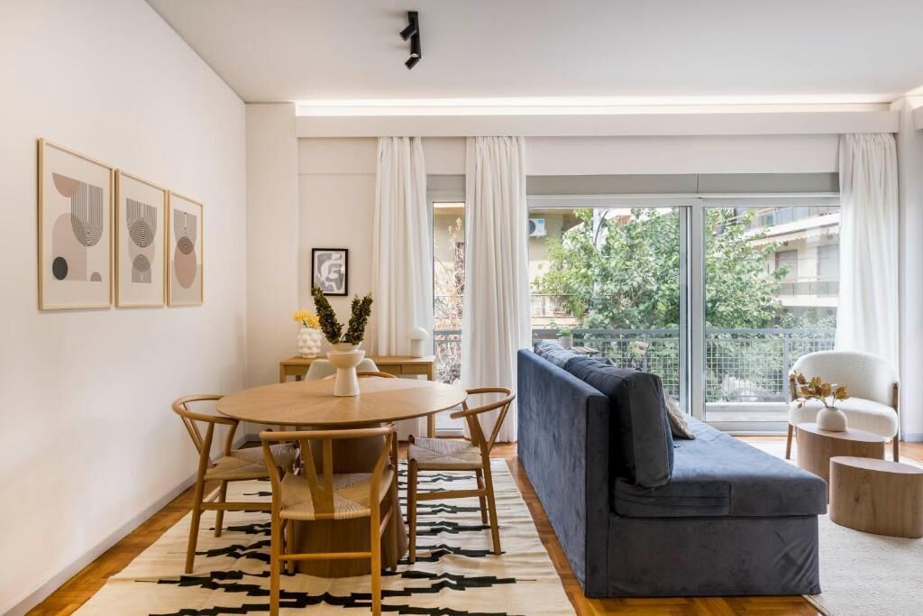 A modern living room with a round wooden table, a grey sofa, and a balcony with a view of greenery.