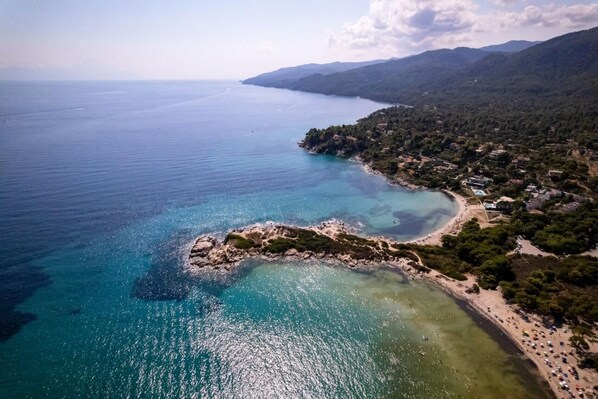 An aerial view of a coastal area with clear waters, a rocky island, and a beach with buildings and greenery.