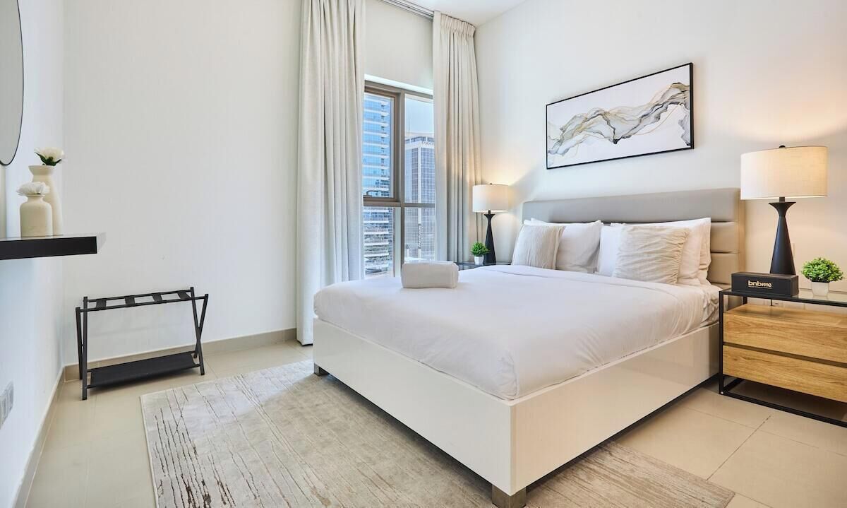 Ein modernes Hotelzimmer mit einem großen Bett, Nachttischlampen, einem kleinen Tisch und Blick auf die Stadt durch das Fenster.