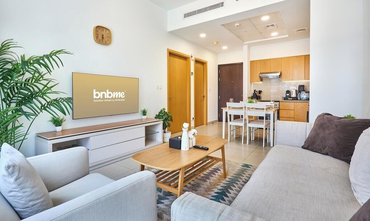 Una sala de estar moderna con sofá, mesa de centro y televisor. Se observa una zona de cocina con armarios y una mesa de comedor.