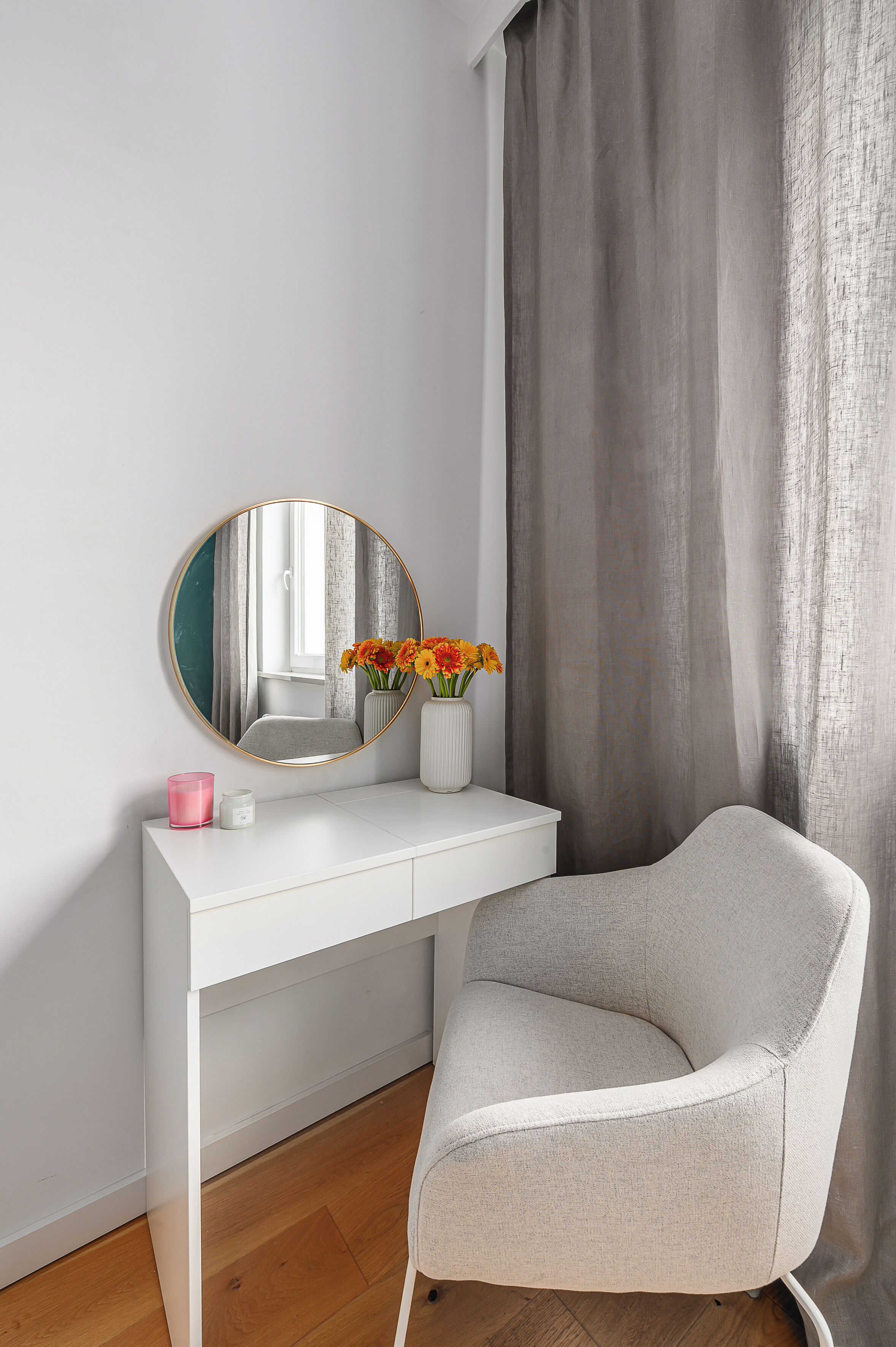 A white vanity with a round mirror, a vase with flowers, and a pink candle.