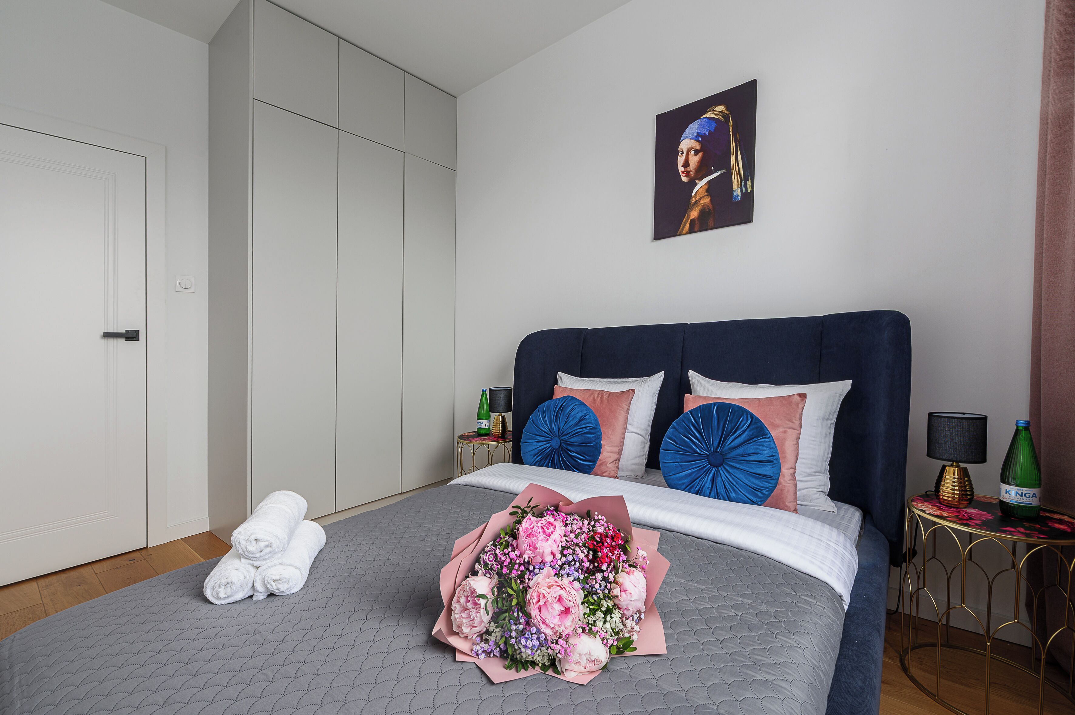 A neatly made bed with a bouquet of flowers, a bottle of champagne, and a painting on the wall.