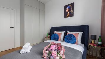 A neatly made bed with a bouquet of flowers, a bottle of champagne, and a painting on the wall.