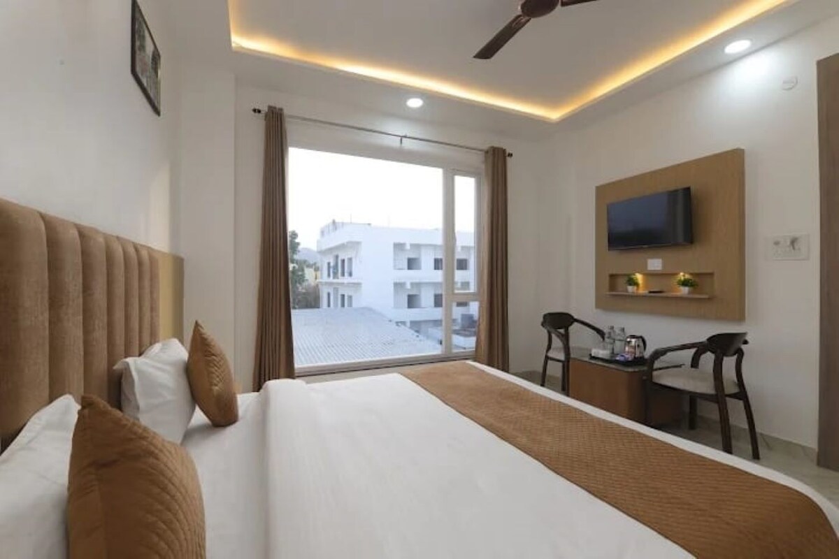 A hotel room with a bed, a desk, a chair, a television, and a view of buildings through a window.