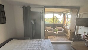 Ein Schlafzimmer mit einem Bett, einem kleinen Tisch und einem Sessel. Ein Glastür öffnet sich zu einem Balkon mit einer Couch und einer Pflanze.
