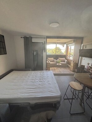 Ein Schlafzimmer mit einem Bett, einem kleinen Tisch und einem Sessel. Ein Glastür öffnet sich zu einem Balkon mit einer Couch und einer Pflanze.