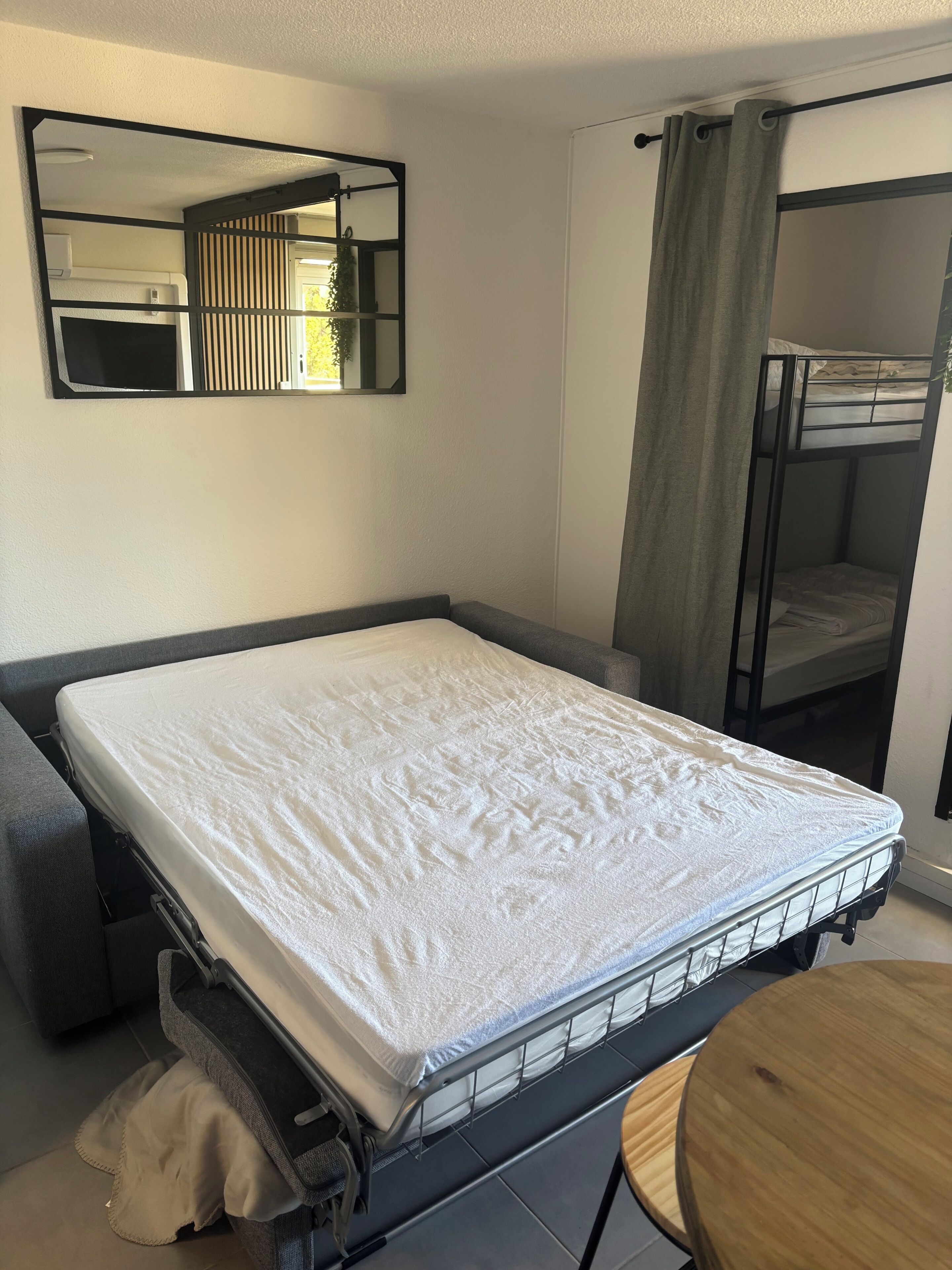 A single bed with a metal frame, a small round table, a mirror, and a bunk bed in the background.