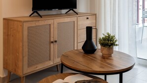 En moderne stue med et fladskærms-tv på et træskab, et rundt træbord med en bog og en vase, samt udsigt til det fri gennem lette gardiner.