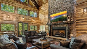 A log cabin interior with a stone fireplace, leather furniture, and large windows. - Splashin Retreat- Indoor Pool, Theater Room, Game Room, Fire Pit (Sevierville)