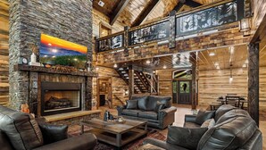 A log cabin interior with a fireplace, leather sofas, a dining area, and a staircase. - Splashin Retreat- Indoor Pool, Theater Room, Game Room, Fire Pit (Sevierville)