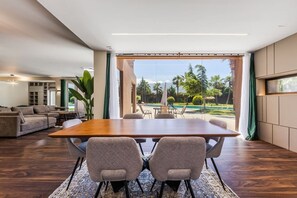 Un comedor moderno con una mesa de madera y cuatro sillas, un sofá, y un ventanal con vistas a palmeras y una piscina.