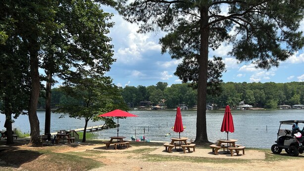 Un espace de pique-nique au bord du lac, avec des tables et des parasols rouges, entouré d’arbres et agrémenté d’un chariot de golf.