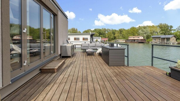 Une terrasse en bois avec des meubles d’extérieur, une pièce fermée par des baies vitrées et une vue sur un lac, avec des bâtiments en arrière-plan.