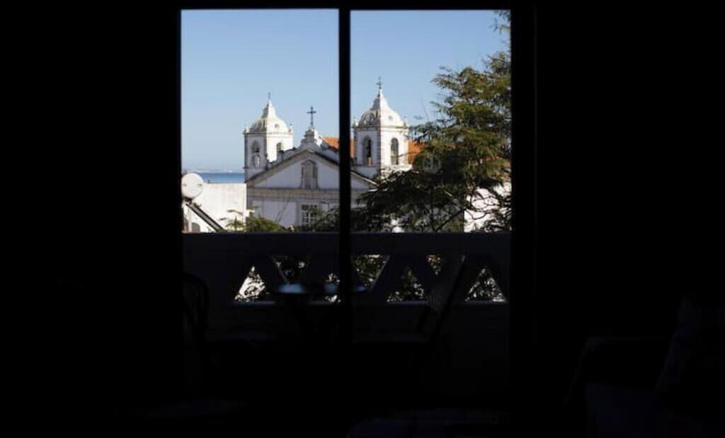 A view through a window with a balcony, featuring a building with two domes and a cross.
