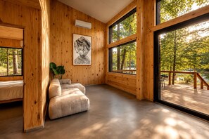 A room with wooden walls, a large window, a bed, a sofa, and a framed artwork. - Serene Modern Tiny Home with Fiery Gizzard Reservoir Views Near Tracy City | Tennessee (Tracy City)