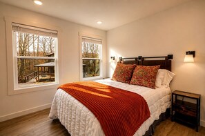 A bedroom with a large bed, two windows with views of a park, a nightstand, and a wall-mounted lamp. - Scenic Bluffside Tiny Home Retreat with Kid-Friendly Loft near South Cumberland in Monteagle, Tennessee (Monteagle)