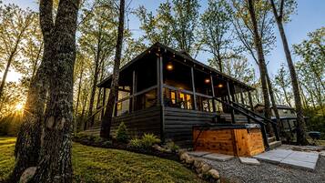 A cabin with a deck, surrounded by trees, and a gravel path leading to the entrance.
