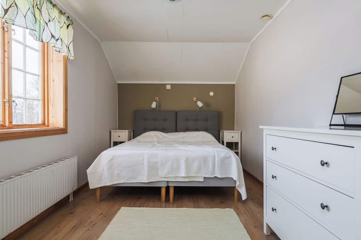 Un dormitorio con cama, mesitas de noche, cómoda, radiador y ventana con cortinas.