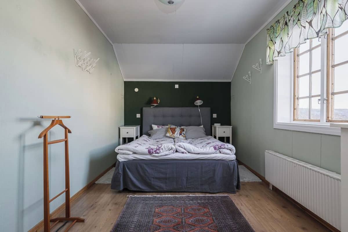 Un dormitorio con cama, mesitas de noche, un radiador, una ventana con cortinas y una lámpara colgante.