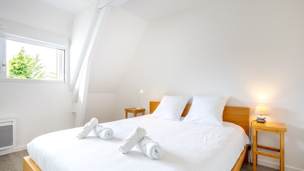 Un lit bien fait, avec du linge de lit blanc et des serviettes pliées, une lampe de chevet et une fenêtre donnant sur un paysage verdoyant.