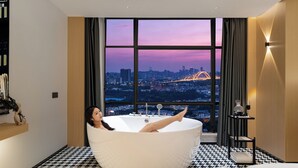 Una mujer se relaja en una bañera grande con vista a un paisaje urbano a través de un ventanal.