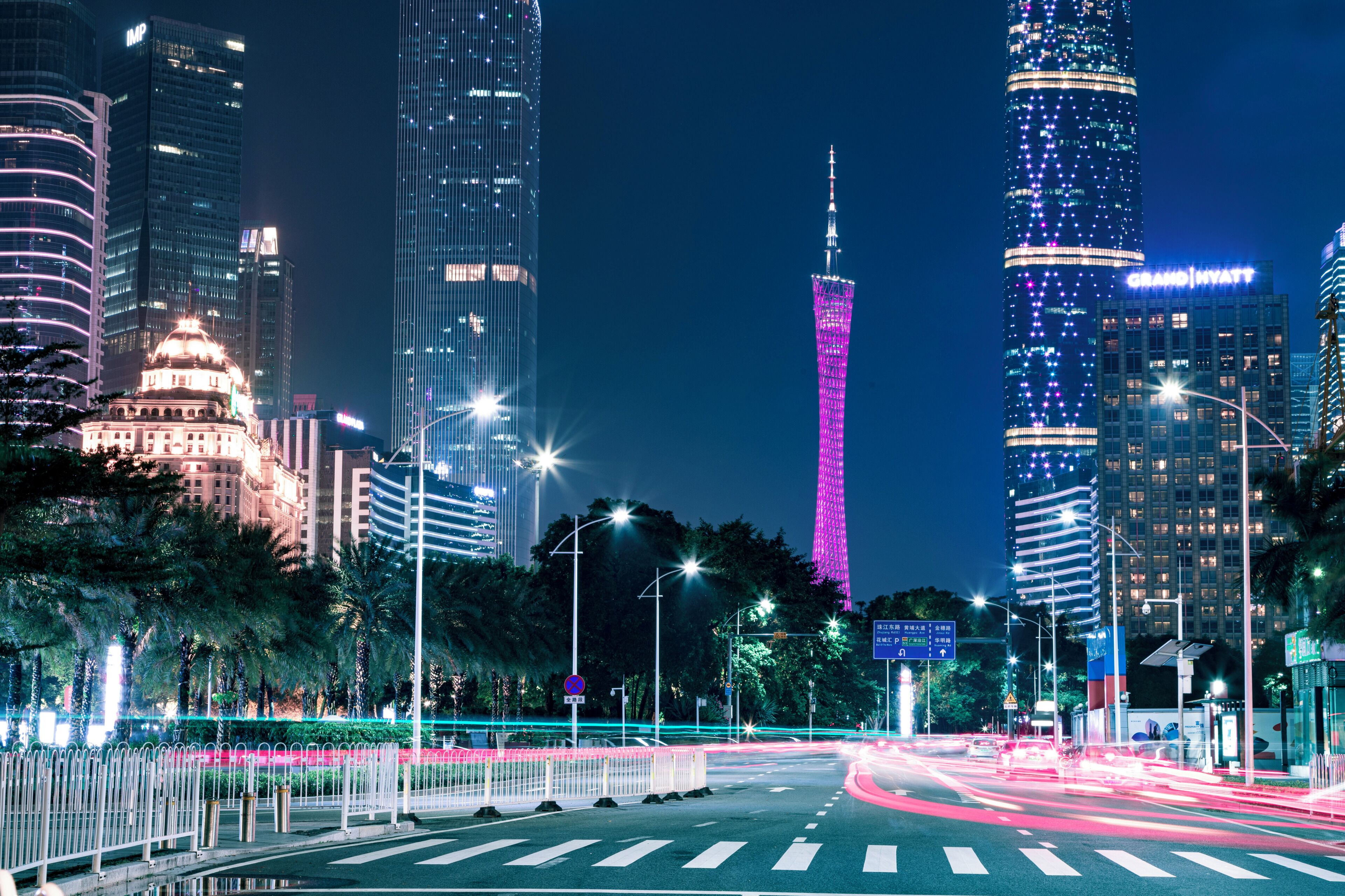 Un paisaje urbano con rascacielos iluminados, una torre destacada con luces rosadas y una intersección bien iluminada con semáforos.