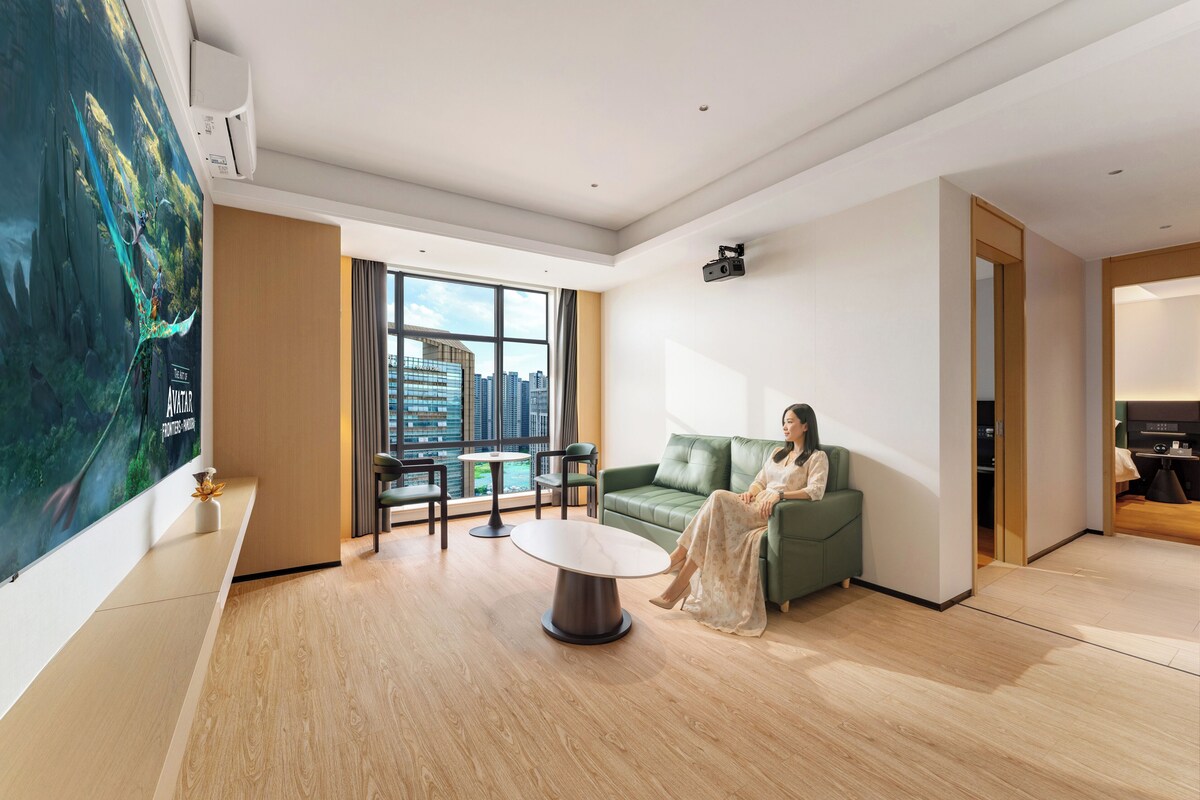 Quarto de hotel moderno com uma janela grande, um sofá verde, uma mesa redonda e um quadro na parede.