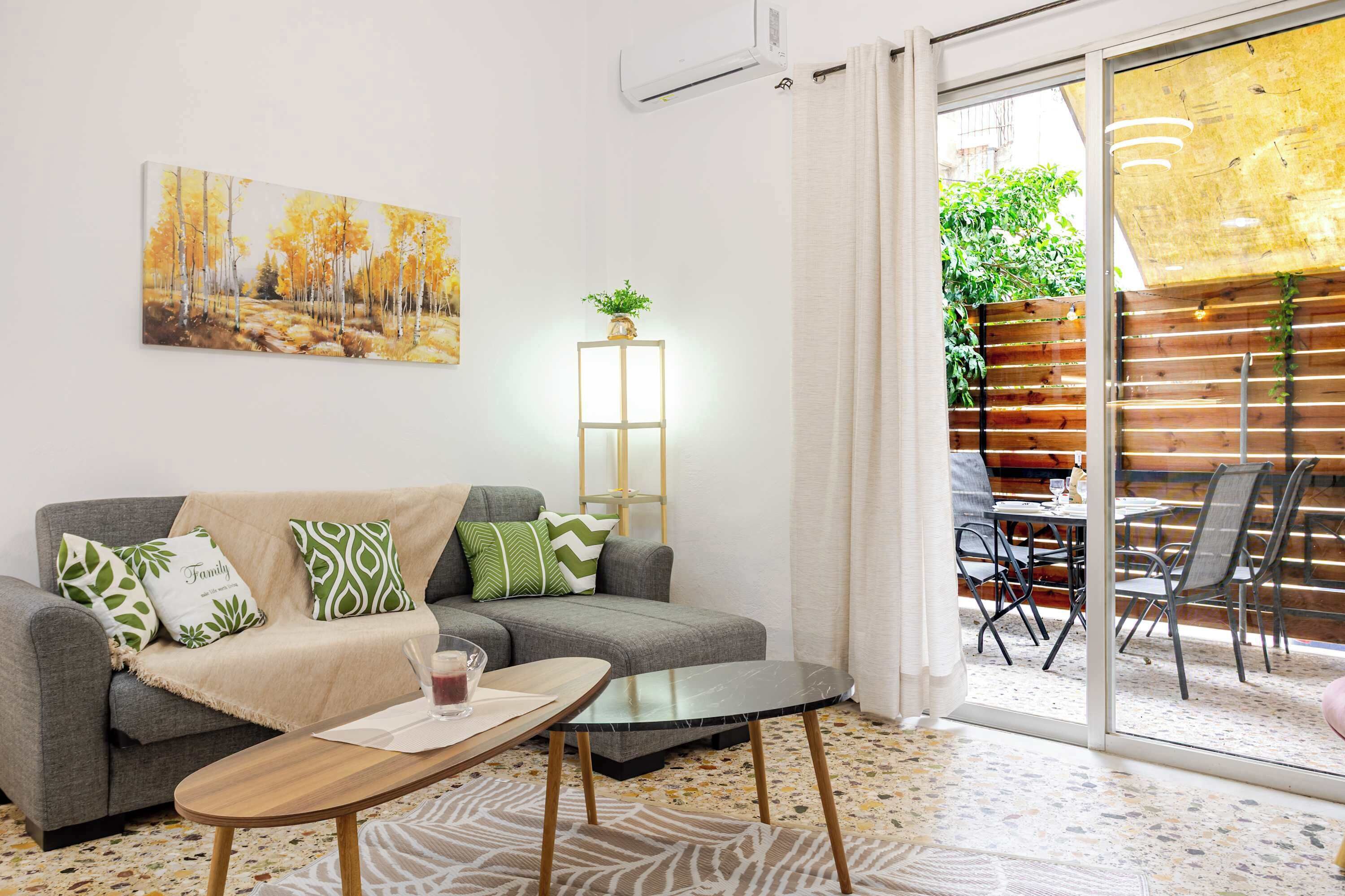 A living room with a sofa, a coffee table, a painting, a plant, and a sliding glass door leading to an outdoor area.
