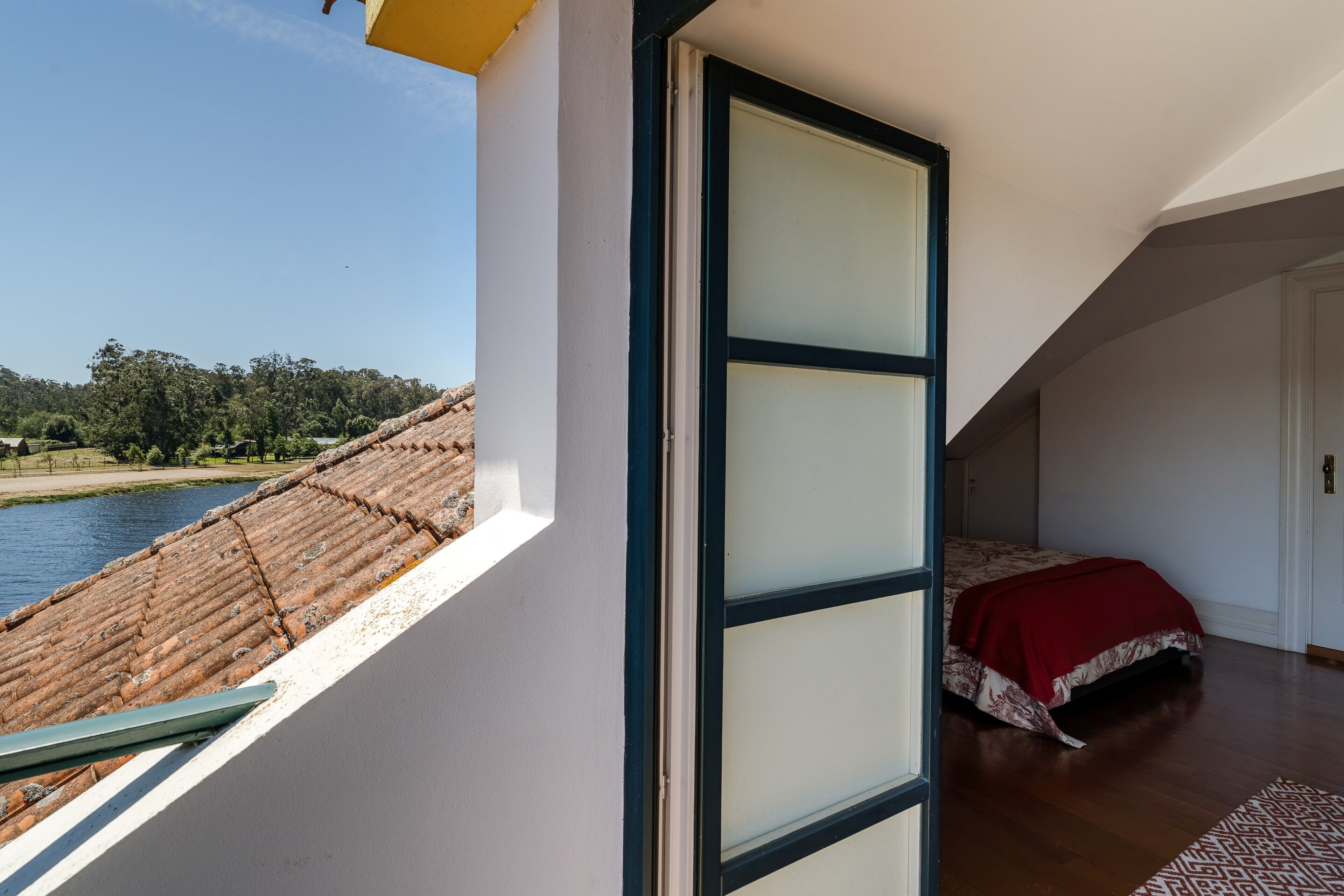 A bedroom with a view of a river, a bed with a red blanket, and a window with frosted glass panels.