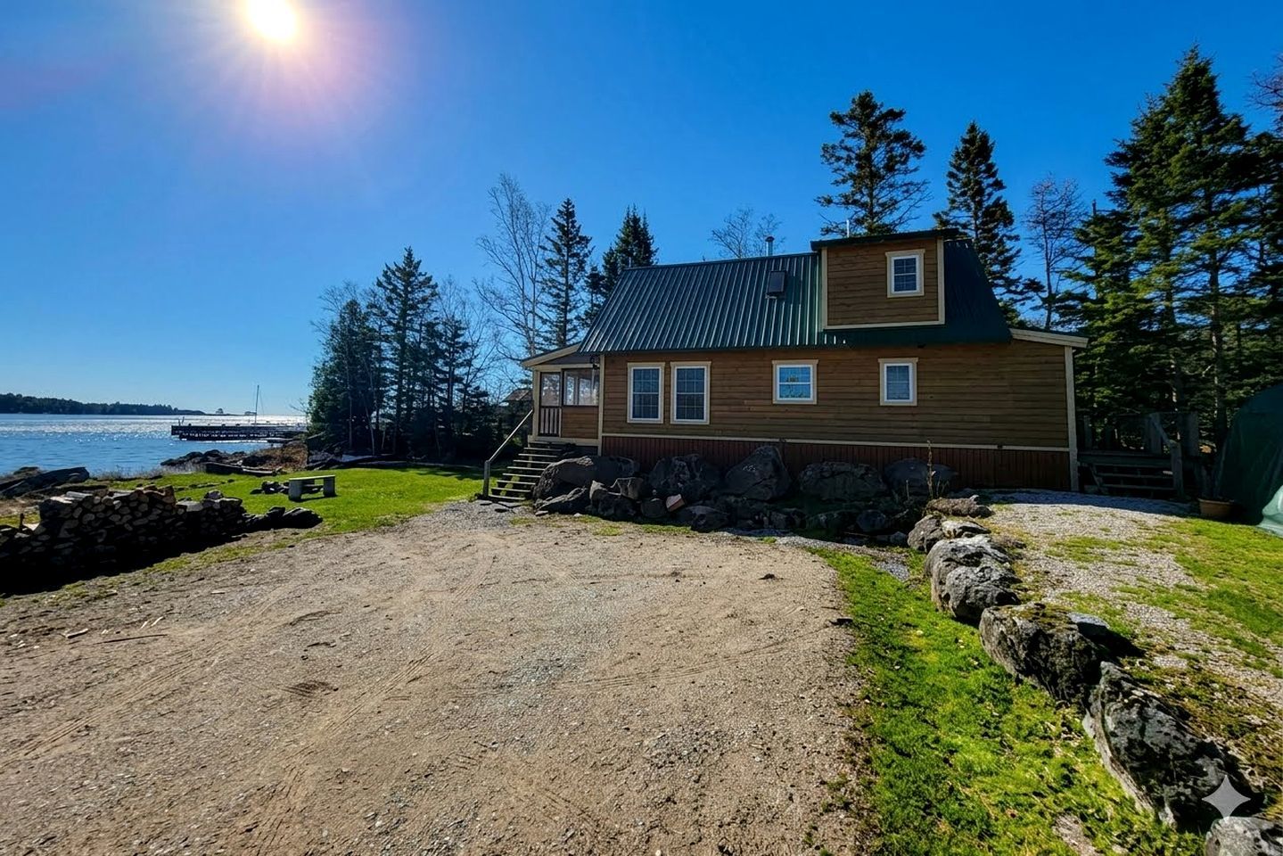 A wooden cabin with a metal roof, a gravel driveway, and a view of the water.