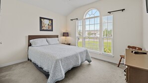 Une chambre à coucher comprenant un lit, un bureau, une chaise, un ventilateur de plafond, une fenêtre avec des stores et un tableau accroché au mur.