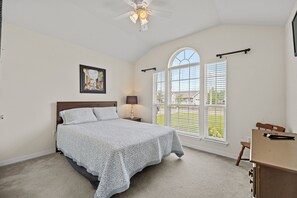 Ein Schlafzimmer mit Bett, Schreibtisch, Stuhl, Deckenventilator, Fenster mit Jalousien und einem Bild an der Wand.