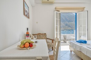 A room with a dining table set with fruits and wine glasses, a bed with towels, a chair, and a view of mountains through an open door. - Katerina's Studio '2' with Sea View and Private Balcony (Arginonta)
